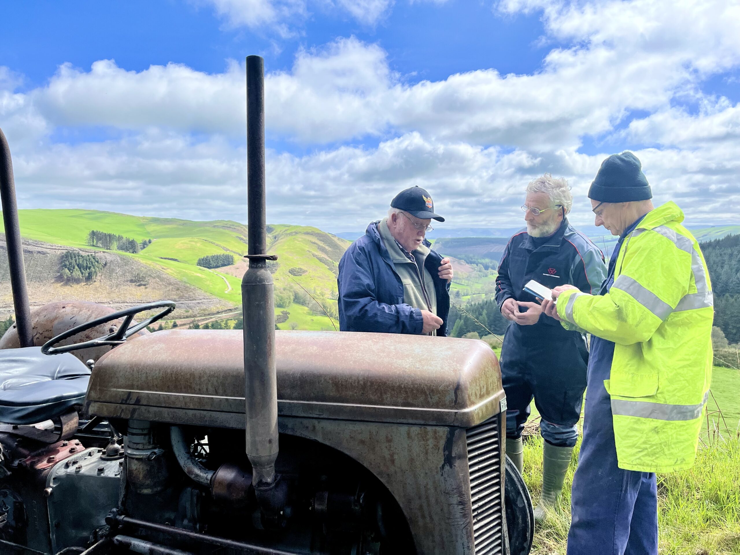 There men talk whilst stood next to old tractor with blue sky clouds and hills in the background.