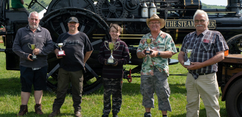 4 adults and a child stood in front of a large steam engine holding winners cups smiling.