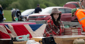 Child wearing fabric hat and a deep red plaid blanked wrapped around them. In the distance the union flag is draped over a table.