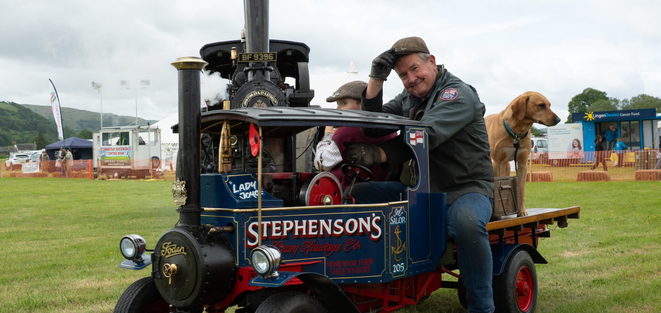 A 1/3 replica of a 1930s Foden C Type steam lorry. With owner riding it and tipping his flat cap to the camera whilst smiling. A young boy and dog sit beside him.